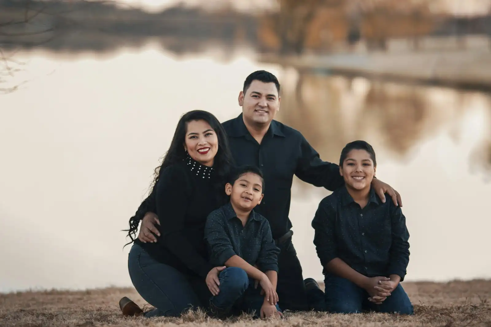A smiling family of four, a mother, a father and two boys, kneeling together by a lake in autumn